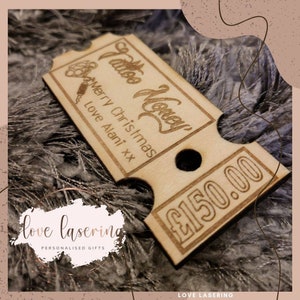 May include: A wooden gift certificate for tattoo money, with the text "Tattoo Money" and "Merry Christmas" in a cursive font. The certificate is decorated with a small tattoo machine image and the text "Love Alani xx". The certificate has a hole punched in the top and the text "&pound;150.00" is printed on the bottom right corner.