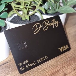 Pu&ograve; includere: Carta di credito in metallo nero con scritte dorate. La carta riporta in alto "D. Bentley" in corsivo, in basso "EXP 12/26 MR DANIEL BENTLEY" e in basso a destra "VISA".
