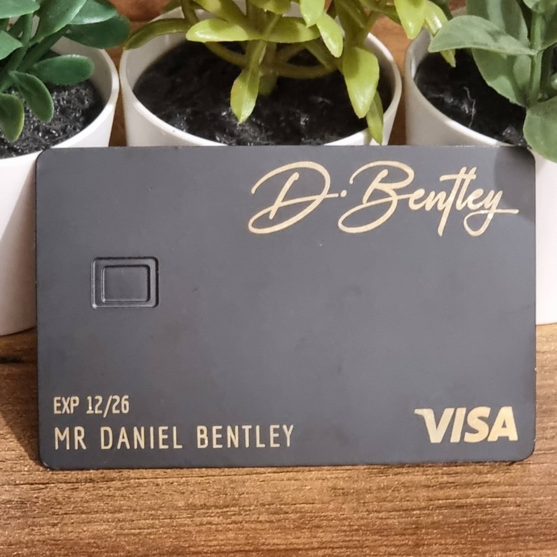 Pu&ograve; includere: Una carta di credito Visa nera con lettere dorate. La carta ha il nome "D. Bentley" e il testo "EXP 12/26 MR DANIEL BENTLEY" stampato su di essa.