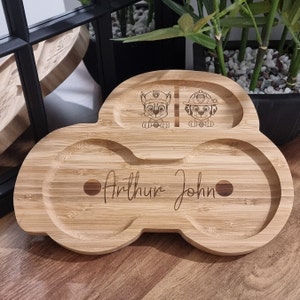 May include: A wooden plate shaped like a car with two separate compartments and a larger compartment in the center. The plate is engraved with the names "Arthur John" and two cartoon characters from the show "Paw Patrol".