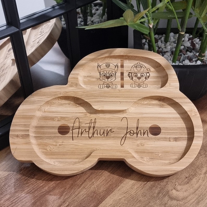 May include: A wooden plate shaped like a car with two separate compartments and a larger compartment in the center. The plate is engraved with the names "Arthur John" and two cartoon characters from the show "Paw Patrol".