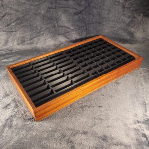 Op de afbeelding: Een rechthoekige houten lade met een raster van kleine, zwarte compartimenten. De lade heeft een warme, roodbruine afwerking en is ontworpen voor het organiseren van kleine items. De binnenkant is bekleed met zwart materiaal, wat een contrast creëert met het hout.