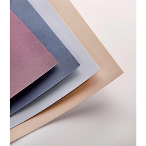 Op de afbeelding: Een stapel getextureerde papieren vellen in tinten mauve, marineblauw, lichtgrijs en beige. De papieren zijn diagonaal gerangschikt, waarbij hun verschillende kleuren en subtiele glans worden getoond. De achtergrond is effen wit.