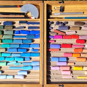 K&ouml;nnte beinhalten: Eine Holzkunstbox gef&uuml;llt mit einer Vielzahl von Pastellkreiden in einem Farbspektrum von Blau- und Gr&uuml;nt&ouml;nen bis hin zu Rot- und Gelbt&ouml;nen. Die Box enth&auml;lt auch Bleistifte, einen Schleifblock und andere Kunstutensilien. Die Anordnung ist organisiert und ansprechend.
