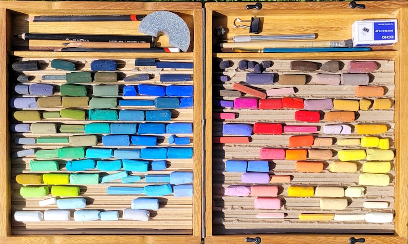 K&ouml;nnte beinhalten: Eine Holzkunstbox gef&uuml;llt mit einer Vielzahl von Pastellkreiden in einem Farbspektrum von Blau- und Gr&uuml;nt&ouml;nen bis hin zu Rot- und Gelbt&ouml;nen. Die Box enth&auml;lt auch Bleistifte, einen Schleifblock und andere Kunstutensilien. Die Anordnung ist organisiert und ansprechend.