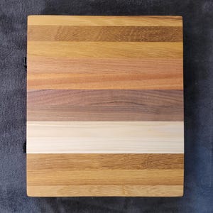K&ouml;nnte beinhalten: Ein rechteckiges Holzschneidebrett mit einer Vielzahl von Holzt&ouml;nen, darunter helle und dunkle Braunt&ouml;ne, Beige und Creme. Das Holz ist in horizontalen Streifen angeordnet. Das Brett hat ein dunkles Metallscharnier auf der linken Seite.