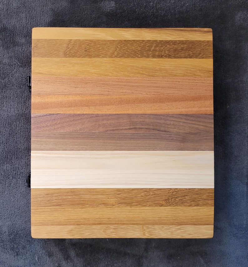 K&ouml;nnte beinhalten: Ein rechteckiges Holzschneidebrett mit einer Vielzahl von Holzt&ouml;nen, darunter helle und dunkle Braunt&ouml;ne, Beige und Creme. Das Holz ist in horizontalen Streifen angeordnet. Das Brett hat ein dunkles Metallscharnier auf der linken Seite.