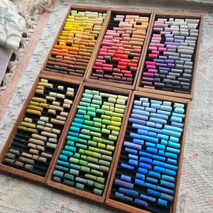 May include: Six wooden boxes filled with rows of colourful pastel chalk sticks. The boxes are arranged in a staggered pattern, with the top row of boxes slightly offset from the bottom row. The chalk sticks are arranged in a rainbow of colours, from red to blue to green.