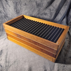 Może przedstawiać: Drewniana skrzynka z czarnym filcowym wyściełaniem i wieloma szczelinami. Skrzynka wykonana jest z trzech różnych rodzajów drewna, tworząc efekt warstwowy.