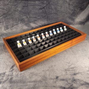 Op de afbeelding: Een houten schilderspalet met een raster van kleine compartimenten, elk met een klein flesje aquarelverf. De lade is gemaakt van lichtbruin hout en heeft een donker interieur.