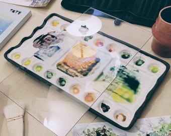 Könnte beinhalten: Eine rechteckige Künstlerpalette mit schwarzem Rand, gefüllt mit verschiedenen Farben. Eine transparente Kunststoffabdeckung liegt obenauf. Ein Pinsel und ein Aquarellgemälde sind im Vordergrund zu sehen. Ein Buch mit dem Wort "TATE" ist ebenfalls sichtbar.