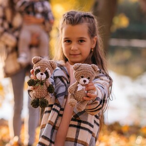 May include: A young girl with long brown hair is holding two crocheted stuffed animals. One is a brown bear with a white bow and the other is a brown and green raccoon. The girl is wearing a plaid shirt and is smiling.