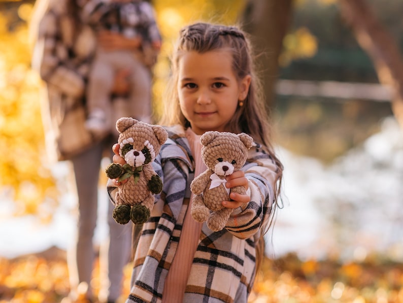 May include: A young girl with long brown hair is holding two crocheted stuffed animals. One is a brown bear with a white bow and the other is a brown and green raccoon. The girl is wearing a plaid shirt and is smiling.
