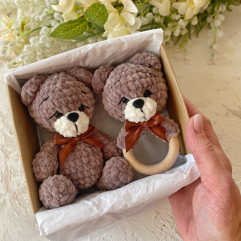 May include: Two plush brown teddy bears with white faces and brown bows are in a gift box with a wooden teething ring.