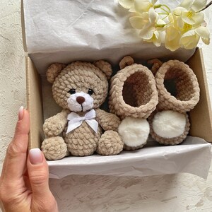 May include: A crocheted brown teddy bear with a white bow sits in a gift box with two pairs of crocheted brown booties with white fur trim.