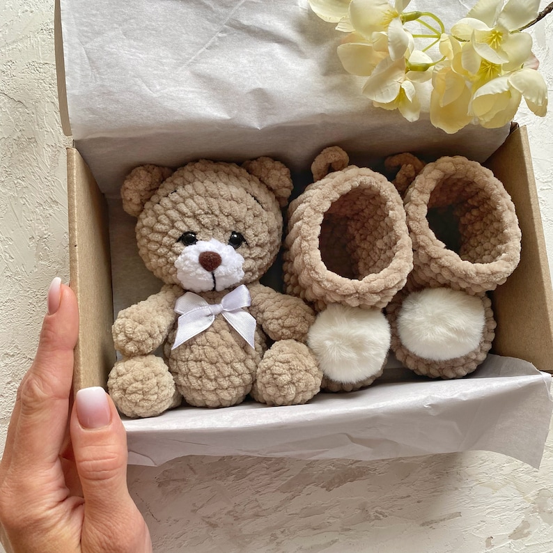 May include: A crocheted brown teddy bear with a white bow sits in a gift box with two pairs of crocheted brown booties with white fur trim.