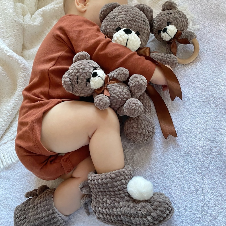 May include: A baby wearing a brown onesie is holding a brown crocheted teddy bear. Two other brown crocheted teddy bears are on the white blanket. One teddy bear has a wooden ring attached to it.