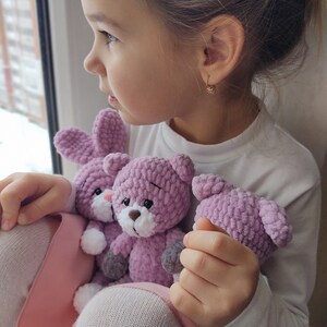 May include: A young girl is holding three crocheted stuffed animals. The animals are a rabbit and two bears. The rabbit is white with pink ears and a pink nose. The bears are pink with white noses and gray paws. The girl is wearing a white shirt and a pink skirt.