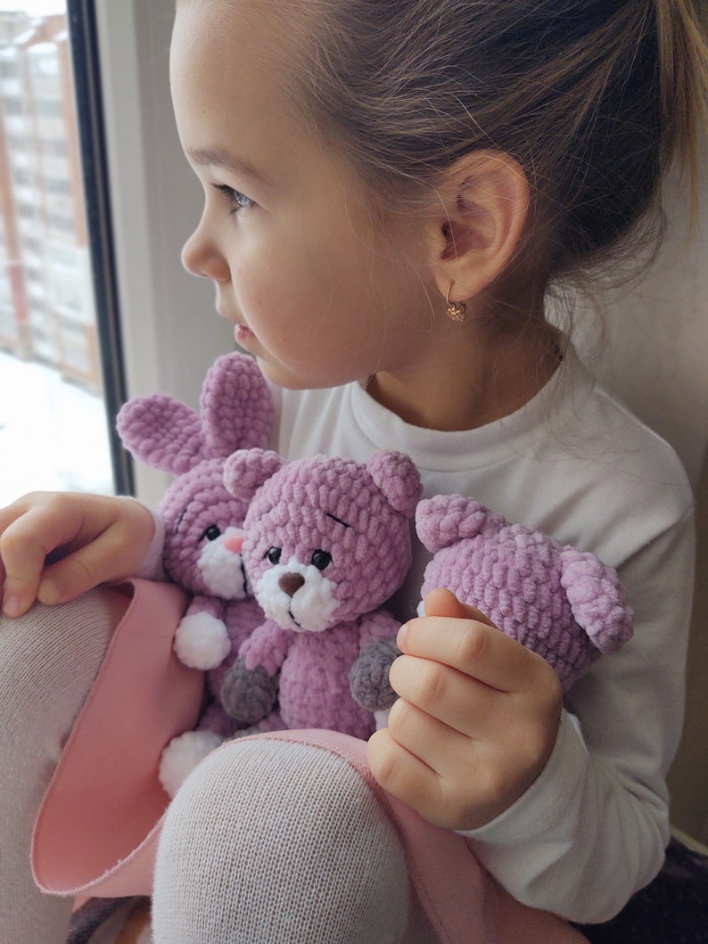 May include: A young girl is holding three crocheted stuffed animals. The animals are a rabbit and two bears. The rabbit is white with pink ears and a pink nose. The bears are pink with white noses and gray paws. The girl is wearing a white shirt and a pink skirt.