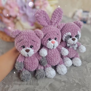 May include: Three crocheted stuffed animals in shades of purple. A bunny, a bear, and a raccoon.