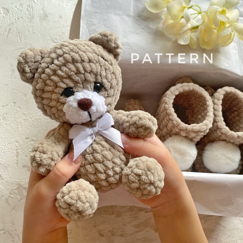 May include: A crocheted teddy bear with a white bow, held in the hands of a person. The bear is light brown and has a white face with black eyes. The word "PATTERN" is visible in the background.
