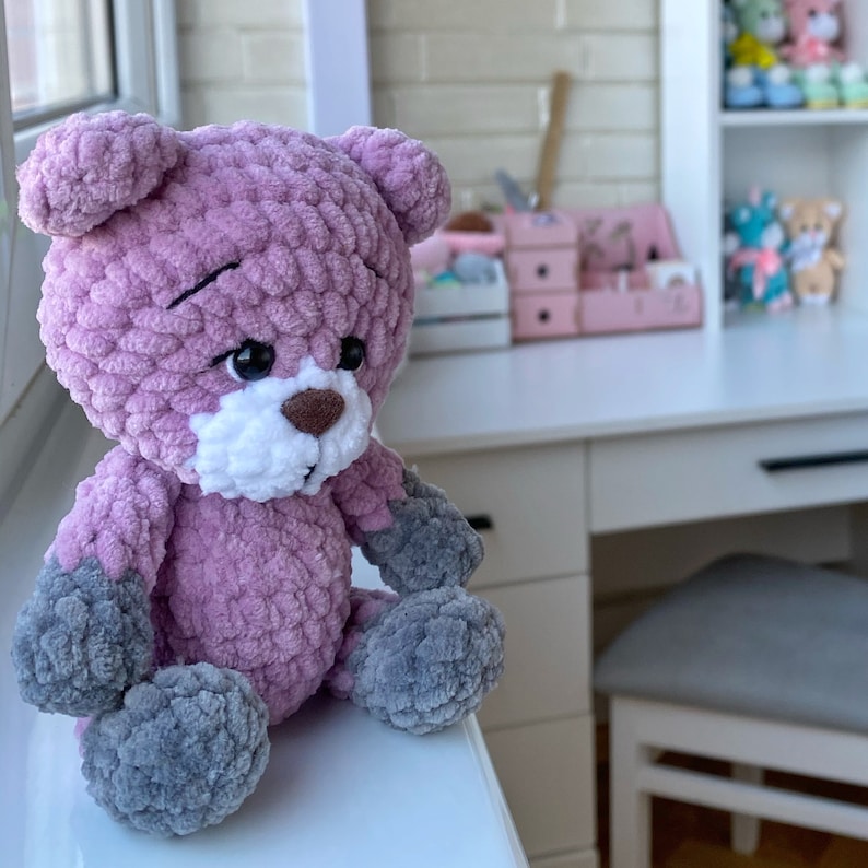 May include: A crocheted plush pink teddy bear with gray accents. The bear has a white nose and black eyes. The bear is sitting on a white surface.