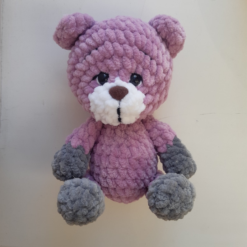 May include: A crocheted plush teddy bear with a purple body, gray arms and legs, and a white face with brown eyes and nose.