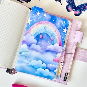 Puede incluir: Una agenda de cuero morado claro con una página cuadriculada, un separador colorido con un arcoíris, nubes y estrellas, tres pegatinas de mariposas, una tarjeta pequeña con contornos de nubes y un bolígrafo de gel rosa.