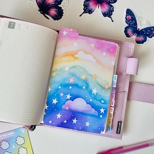 May include: A pink leather-bound planner with a rainbow and stars design on the inside cover. The planner has a tabbed divider system with colorful tabs. The planner is open to a page with a grid pattern. There are several stickers on the cover, including butterflies and a cloud.