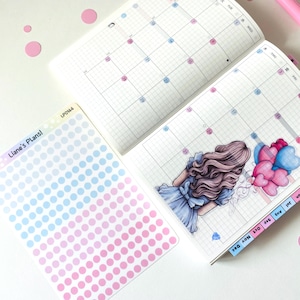 Può includere: Agenda aperto con pagine di calendario, con un disegno di una ragazza con palloncini a forma di cuore. Include adesivi a pois con il testo "Liane's Plans!" e una penna rosa. Schema di colori rosa e blu.