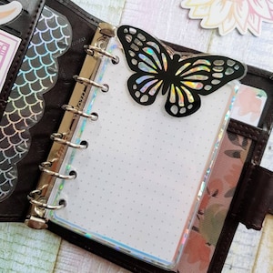 以下が含まれることがあります： 黒い虹色の蝶のステッカーが、黒い革製のバインダーの透明なプラスチックページに貼られています。バインダーはリングバインディングシステムで、白いドットグリッドのページが開かれています。