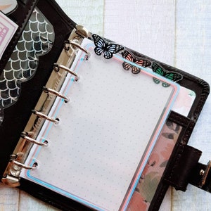 May include: Black leather planner with a clear page protector featuring a dotted grid. The page protector has a pink and blue border. Decorative butterfly paper clips in black, pink, and green adorn the top of the page.