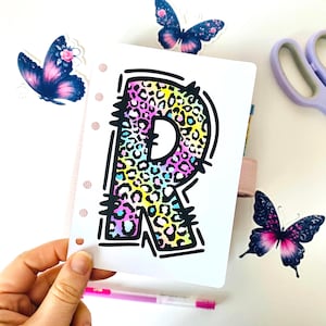Könnte beinhalten: Weißer Planereinsatz mit großem Buchstaben "R" in schwarzer Umrandung und buntem Leopardenmuster. Schmetterlingsaufkleber und ein rosa Stift sind ebenfalls zu sehen.