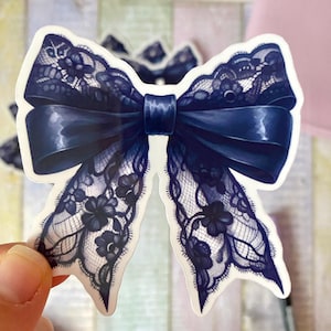 Può includere: Un adesivo a forma di fiocco blu navy con un centro in raso e delicati dettagli in pizzo. L'adesivo presenta un'illustrazione dettagliata di un fiocco con intricati motivi floreali all'interno del pizzo.