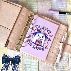 May include: A pink and purple planner with a ghost holding a pumpkin bucket with candy corn and stars. The text on the planner says "Too Cute To Spook".