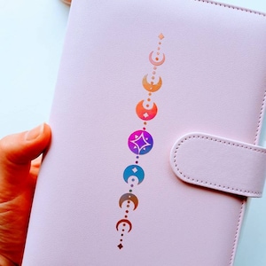 Può includere: Un'agenda in pelle rosa con un design verticale di lune crescenti e una stella in colori arcobaleno.