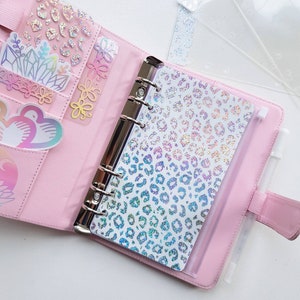 May include: A light pink six-ring binder featuring a holographic leopard print dashboard, multiple interior pockets containing various holographic stickers, and a white pen held in an elastic loop on the side, with clear plastic dividers nearby.