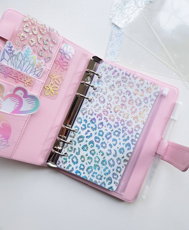 May include: A light pink six-ring binder featuring a holographic leopard print dashboard, multiple interior pockets containing various holographic stickers, and a white pen held in an elastic loop on the side, with clear plastic dividers nearby.