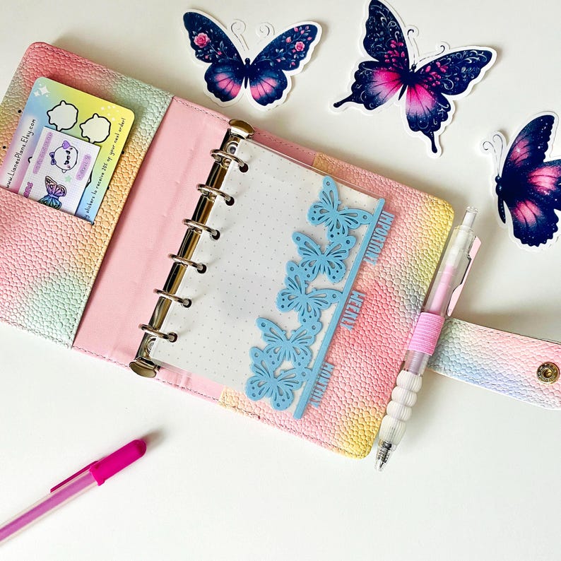 May include: A pastel-colored planner with a pink interior, open to a dotted page. Light blue butterfly cutouts mark the sections. A pink pen and butterfly stickers are also present. Decorative butterfly stickers are scattered around the planner.