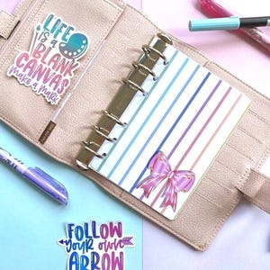 May include: A beige textured planner with a disc-bound insert featuring blue and purple vertical stripes and a pink bow graphic. A sticker reads Life is a blank canvas make a mark and another reads Follow your own arrow. Two pens are visible.