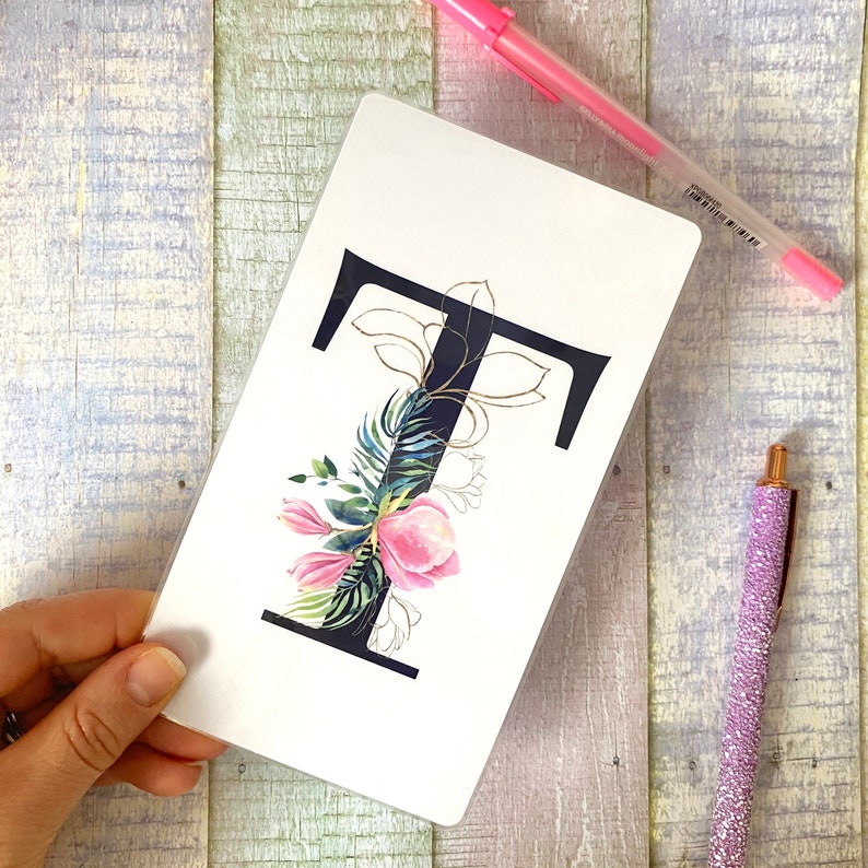 May include: A white notebook with a black letter "T" decorated with pink flowers and green leaves. The letter "T" is outlined in gold.