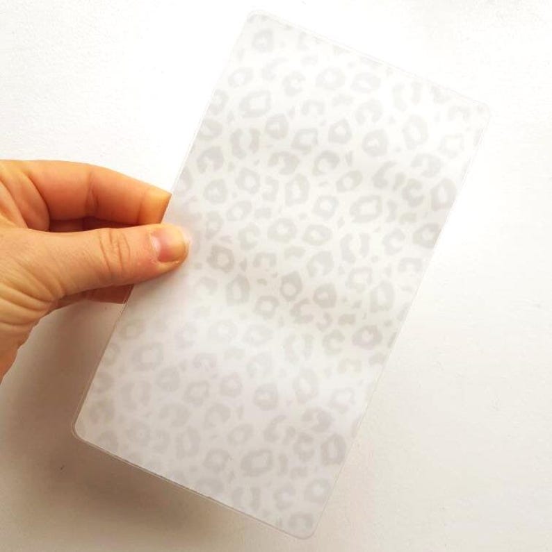May include: A rectangular, semi-transparent plastic embossing folder featuring a repeating, subtle white leopard print pattern across its entire surface, designed for use in crafting machines to create textured impressions on paper or cardstock.