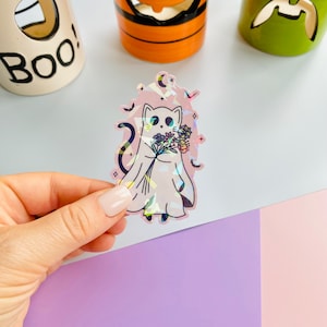 Puede incluir: Una pegatina holográfica con un gato blanco vestido de fantasma que sostiene un ramo de flores. La pegatina tiene un fondo rosa con un efecto brillante.