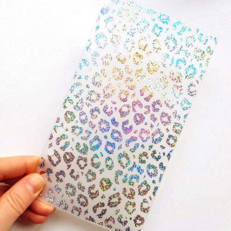 May include: A rectangular translucent plastic sheet featuring a repeating holographic leopard print pattern. The design consists of numerous small, iridescent spots that reflect various colors when light hits the surface of the material.