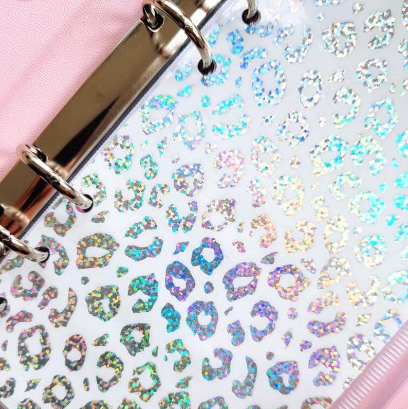 May include: A clear plastic binder divider featuring a repeating holographic leopard print pattern. The divider includes a silver-toned metal six-ring binding mechanism along the left edge and is set against a soft pink background.