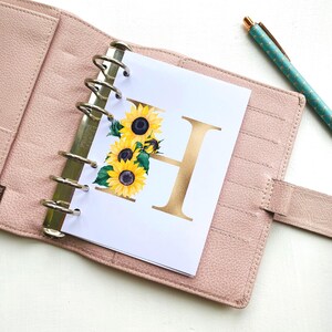 May include: A light pink leather binder with a six-ring metal mechanism holding a white page featuring a large gold letter H decorated with yellow sunflowers and green leaves, accompanied by a teal pen with a gold geometric pattern.