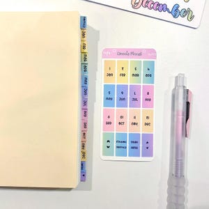 May include: A planner with monthly tabs and a sticker sheet featuring months and numbers. A clear pen with pink ink rests on the white surface. The tabs are labeled with the months of the year.