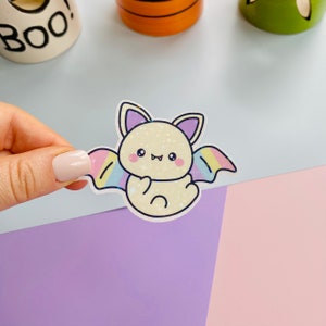 Könnte beinhalten: Eine süße Cartoon-Fledermaus mit Regenbogenflügeln. Die Fledermaus ist gelb mit einer rosa Nase und lila Ohren. Die Flügel sind ein Farbverlauf von rosa, gelb, blau und grün.