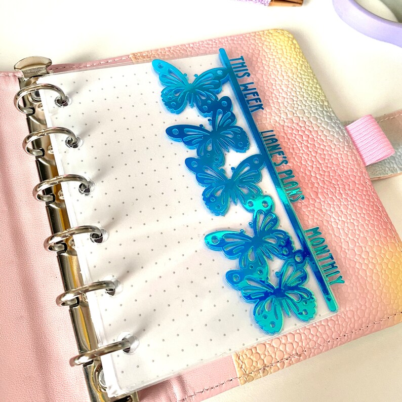 May include: A planner insert with a row of blue butterfly cutouts. The insert is placed inside a pink and rainbow-colored planner. Text on the insert reads "THIS WEEK", "LIANE'S PLANS", and "MONTHLY".