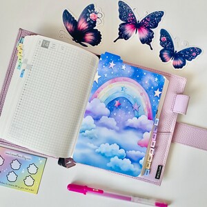 May include: A pink leather planner with a rainbow and stars design on the left page. The right page is a gridded planner with the number 14 at the top. There are several butterfly stickers on the cover of the planner.
